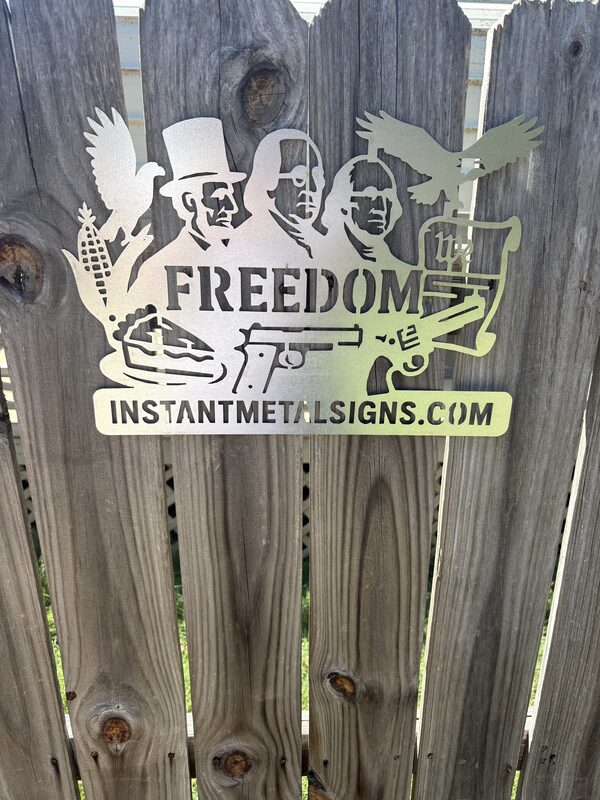 Photo of a laser-cut metal sign reading Freedom, with portraits of historic American figures, eagles, corn, apple pie and pistols, above instantmetalsigns.com, hung on a wooden fence
